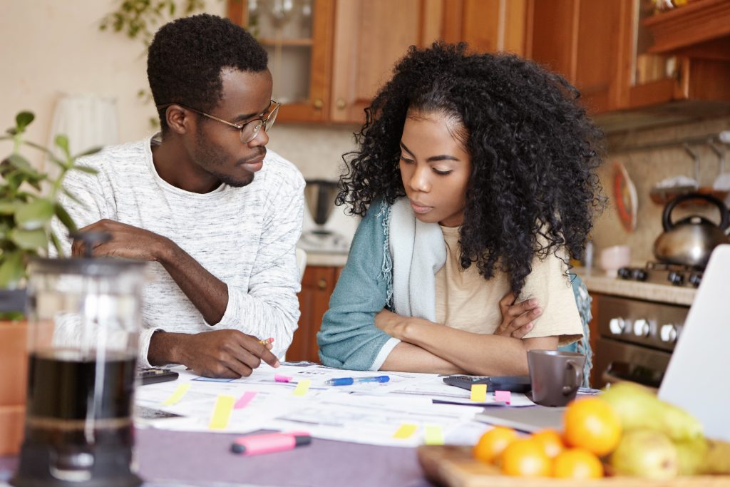 young african couple quarreling because of many debts, sitting at kitchen table with documents, calculating their domestic expenses. wife is mad at her unemployed husband not able to pay bills